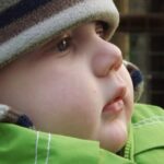 A close up of a baby's face, he is wearing a brown striped hat and a green coat