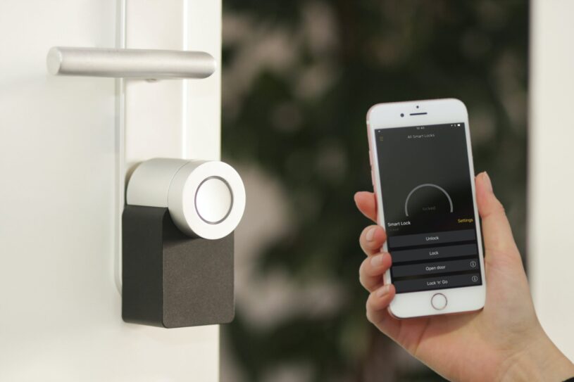 A hand is holding a cell phone with security features on, in front of an external door with a smart lock; Is It Worth Making Your Home Smarter