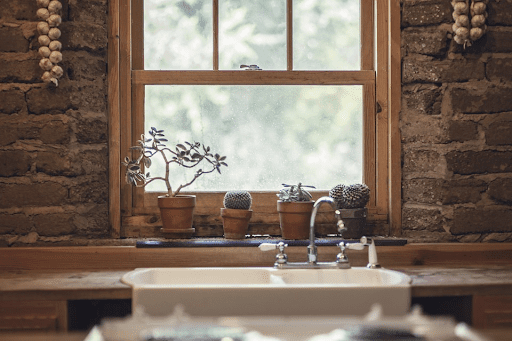 a simple view of a kitchen window. In front of this is a large white sink and four house plants on the window sill; Treating Yourself To A Nicer Kitchen