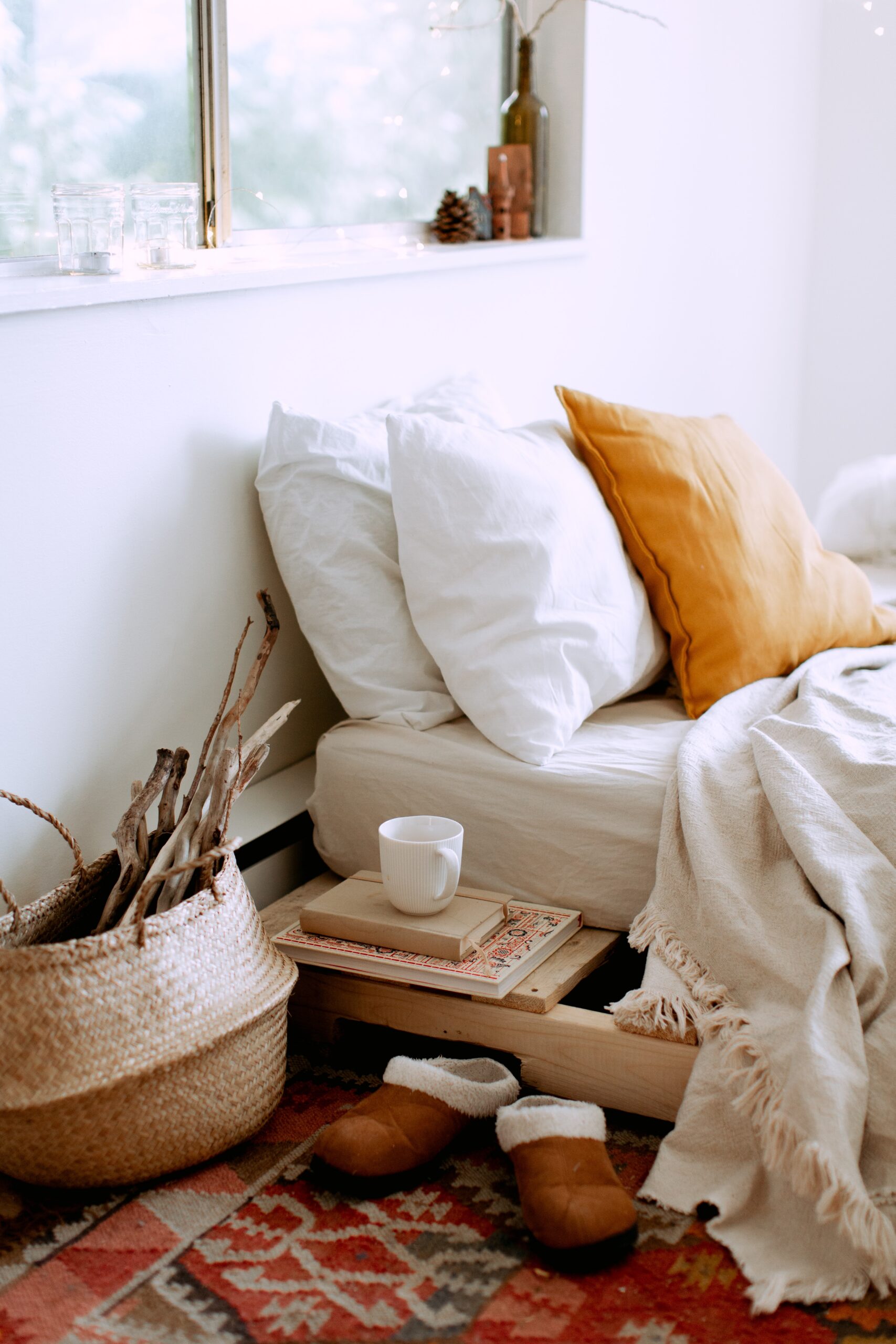 the head end of a bed with 3 pillows and a blanket pulled back, with a seagrass basket, a mug and a pair of slippers on the floor; How To Keep Your Home Warm Without Spending Lots Of Money