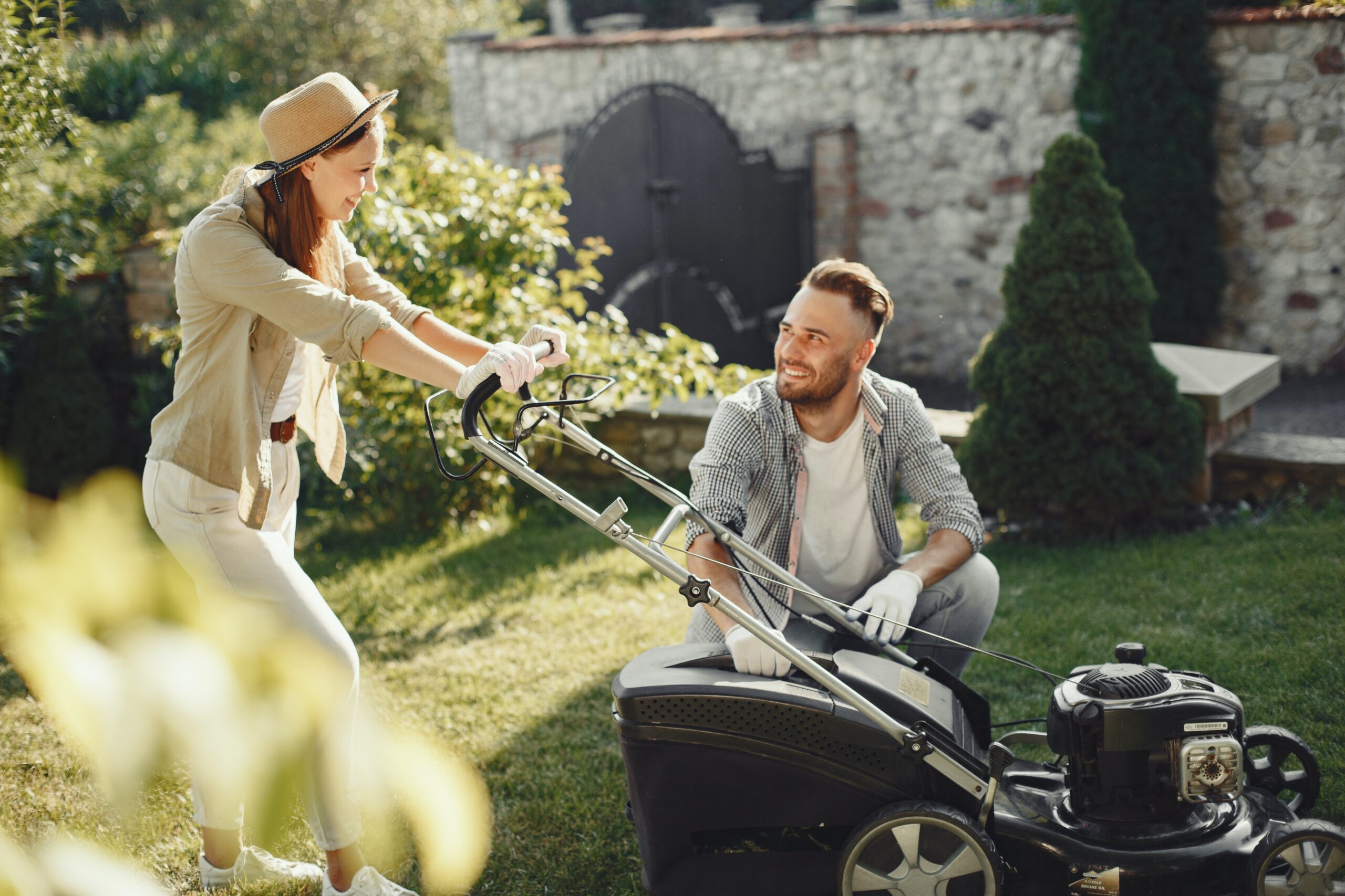 smiling woman in a long sleeve light brown shirt holding onto a lawnmower, with a smiling man knelt down next to it