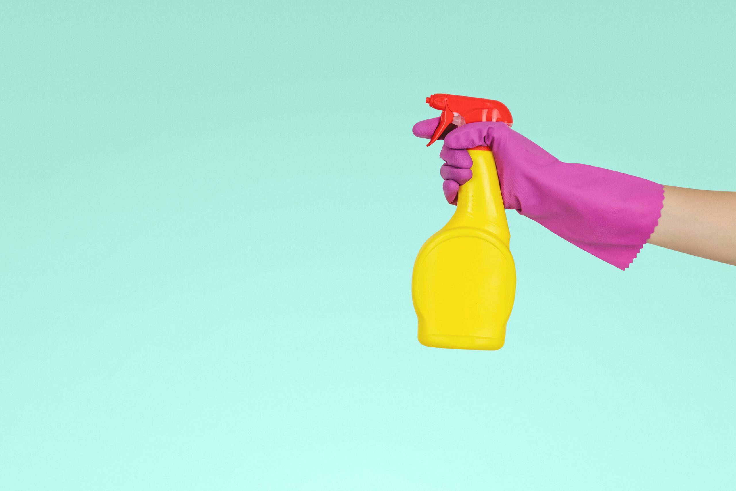a pink gloved hand holding a yellow spray bottle with a red top, against a light blue background; 5 Tips for Spring Cleaning Your Home