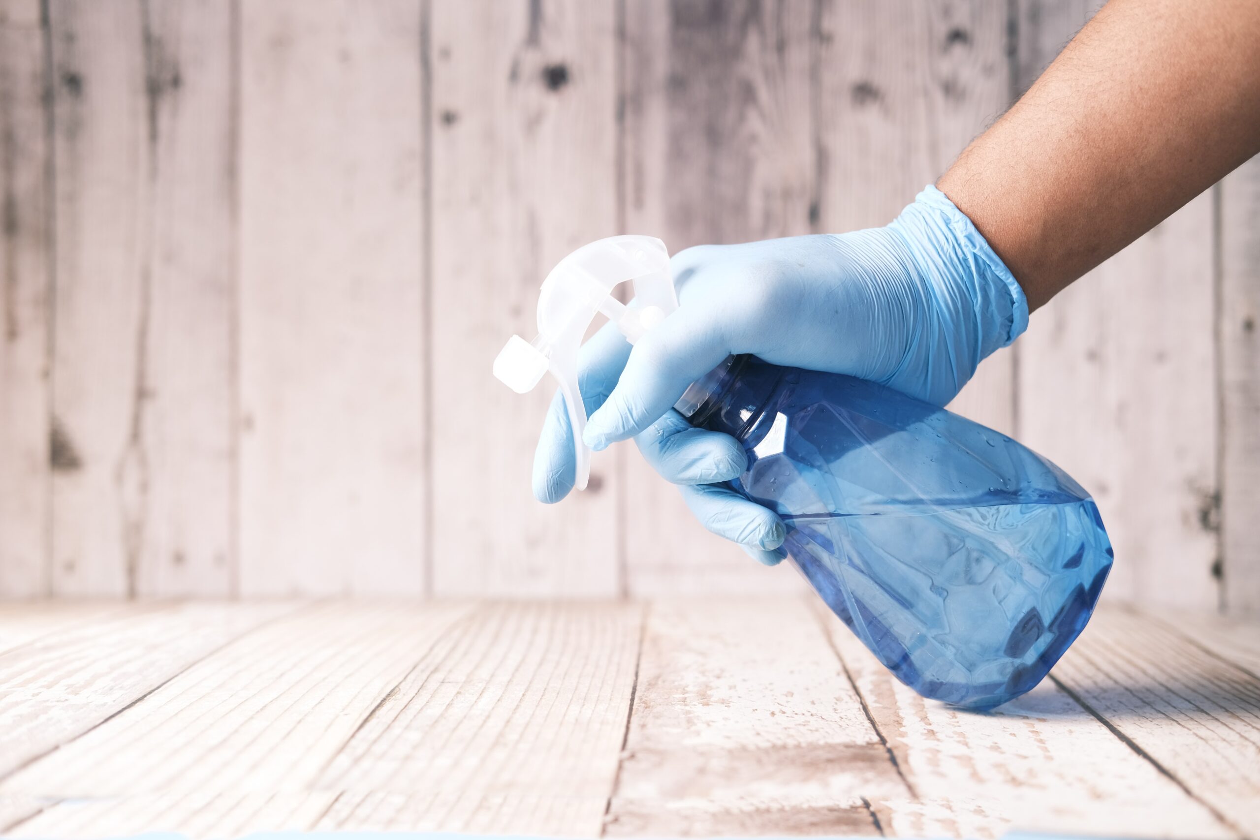 a hand with a blue glove holding a spray bottle above a wooden floor; The Best Ways to Tackle Your Big House Clean This Season