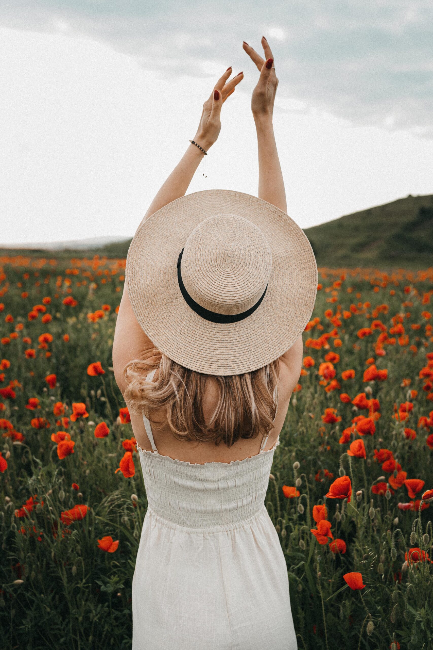 a blonde haired woman wearing a white sleeveless dress and a straw hat with a black trim, standing with her arms in the air in front a field of poppies; Calmness and Clarity How To Be Better at Making Big Decisions
