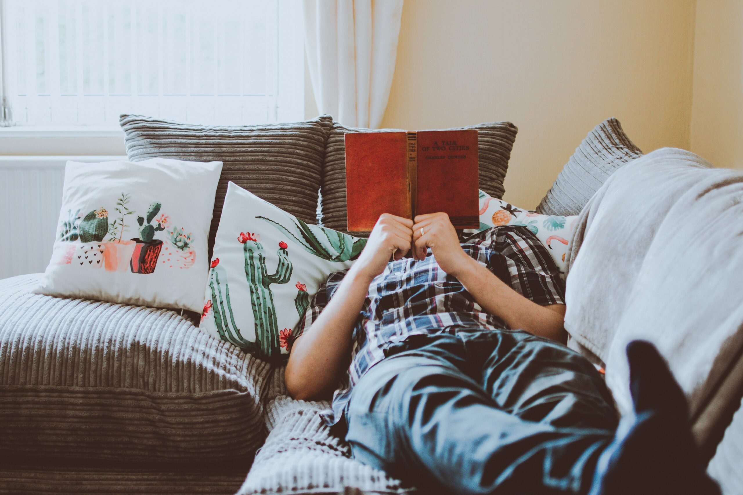 a person laying on a sofa reading a book. The book is covering their face. The cushions on the sofa have pictures of cacti on them