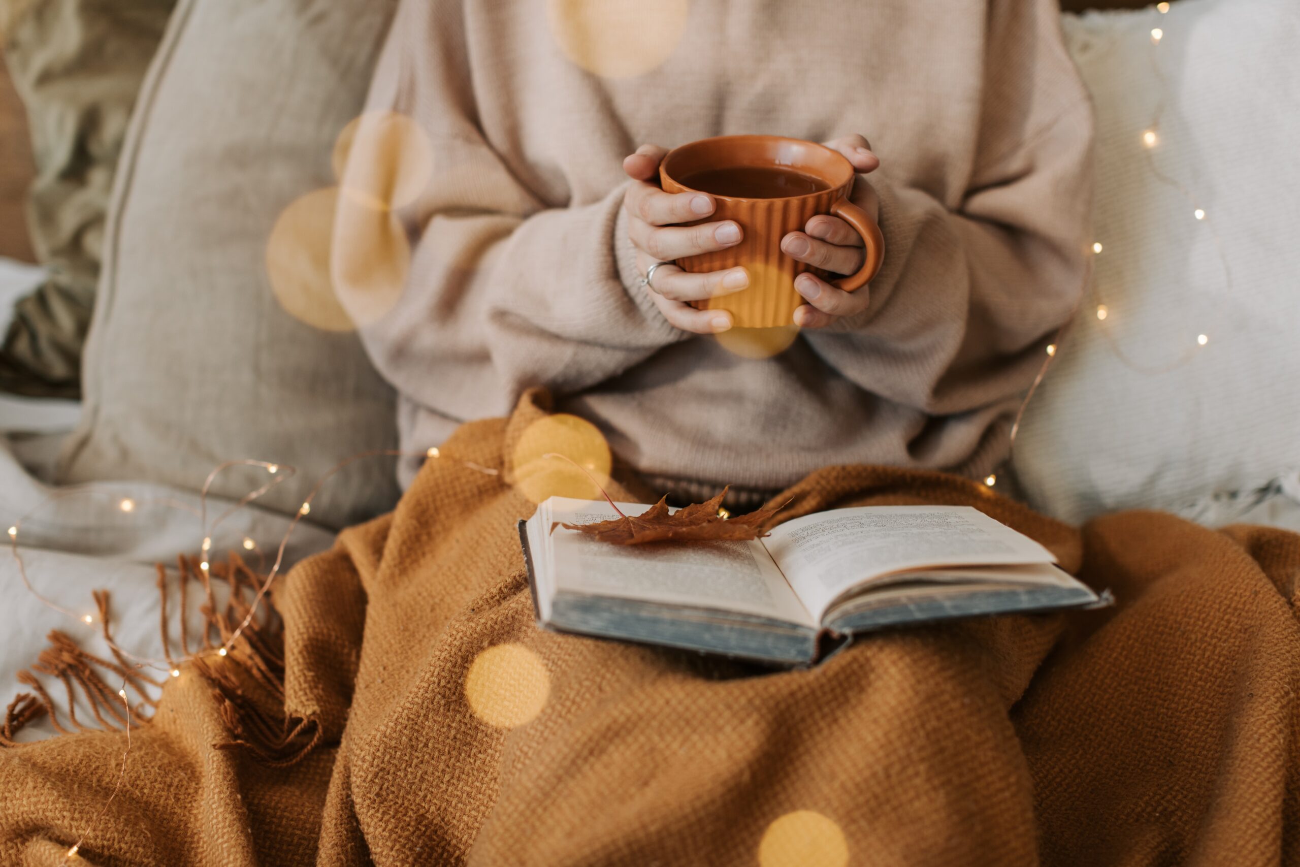 a person in a grey sweater sat under a brown blanket holding a mug with an open book on their knee; 3 Easy Ways To Upgrade The Comfort Of Your Home 