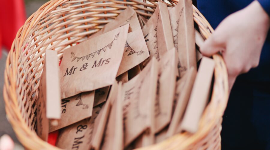 A hand holding a wicker basket containing brown envelopes, with the words Mr & Mrs printed on them. They are filled with wedding confetti