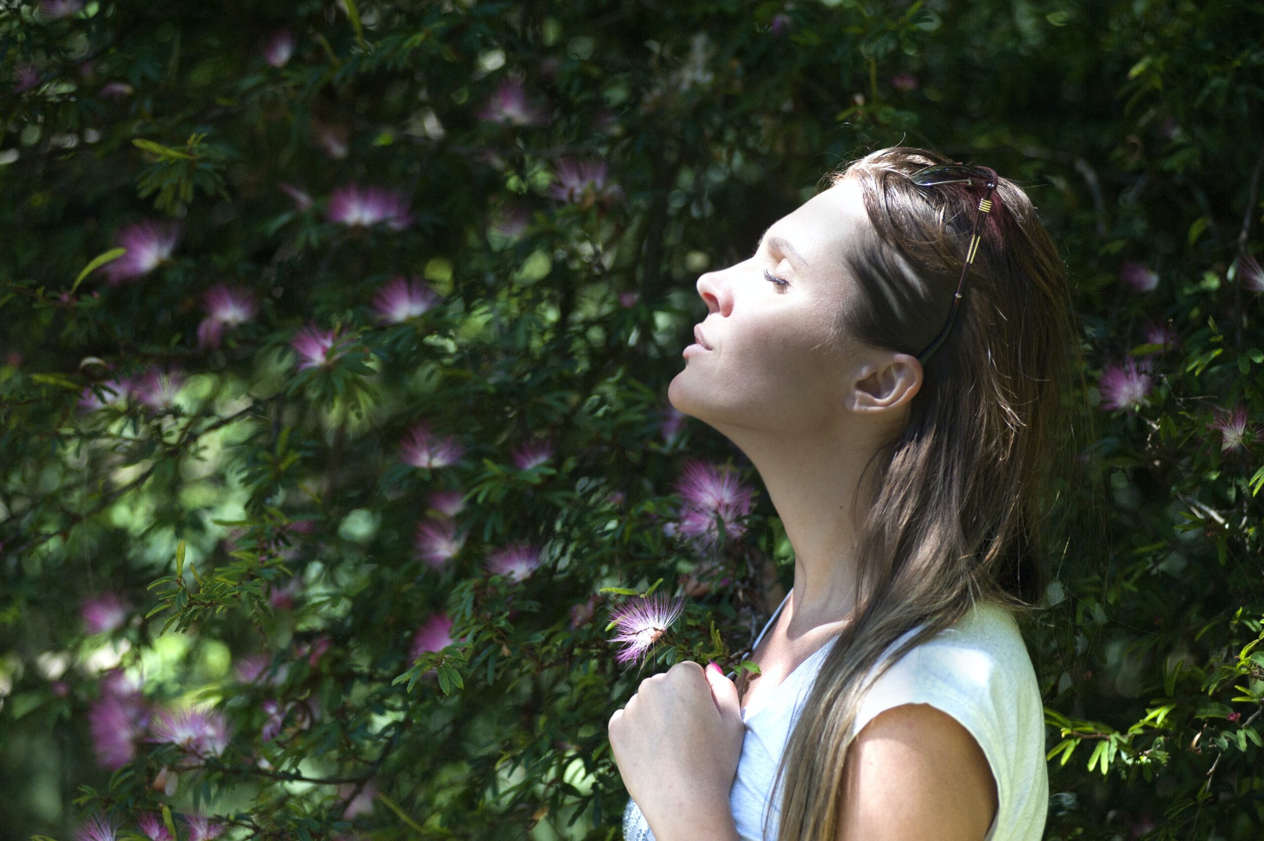 the profile view of a woman with her eyes closed and head raised, stood in front of a green hedge with purple flowers; Getting A Little More Use Out Of Your Garden