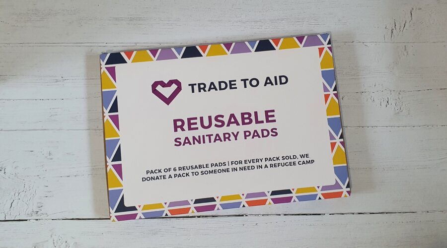 A colourful patterned rectangle box on a white wood effect background. On the box it says "Trade To Aid Reusable Sanitary Pads"