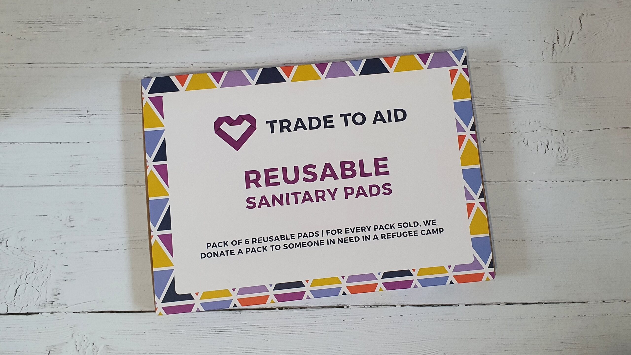 A colourful patterned rectangle box on a white wood effect background. On the box it says "Trade To Aid Reusable Sanitary Pads"