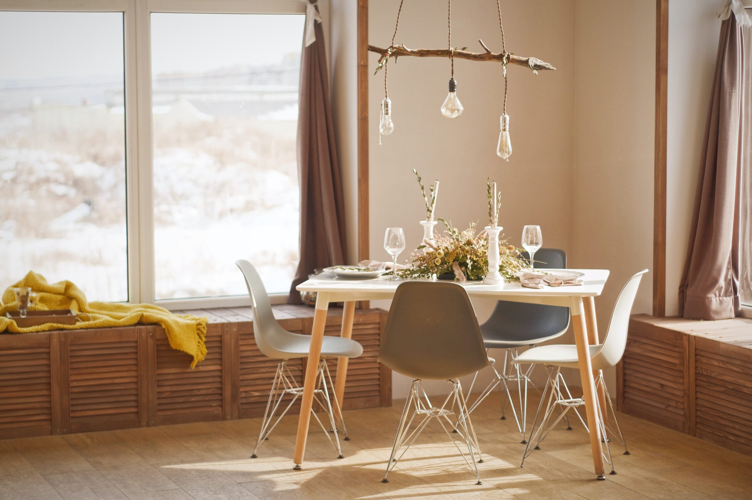 A white table with wooden legs in a light room during daytime. There is a floral centrepiece in the middle of the table. Above the table is a rustic light hanging made from a wooden branch; Rustic Charm Incorporating Rustic Elements into Your Home Interiors