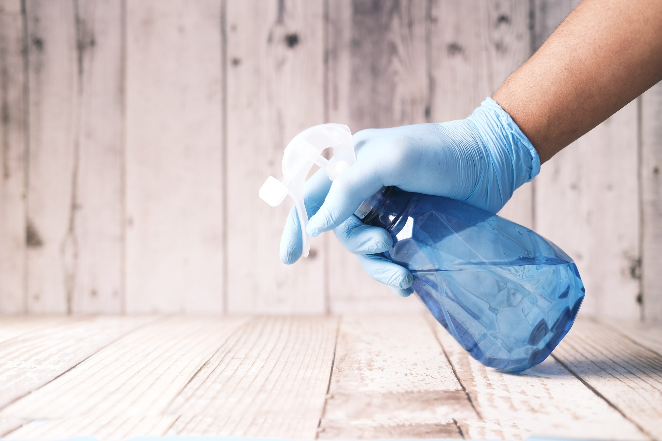 a hand with a a plastic glove holding a cleaning bottle above a wooden surface; Stop Ignoring These Important Cleaning & Maintenance Tasks In Your Kitchen