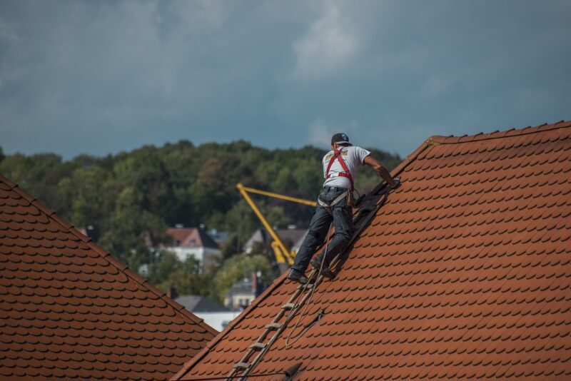 a man on ladders on a house roof repairing tiles. There are green trees in the background and a bright blue sky; 4 Ways To Create A Waterproof Home