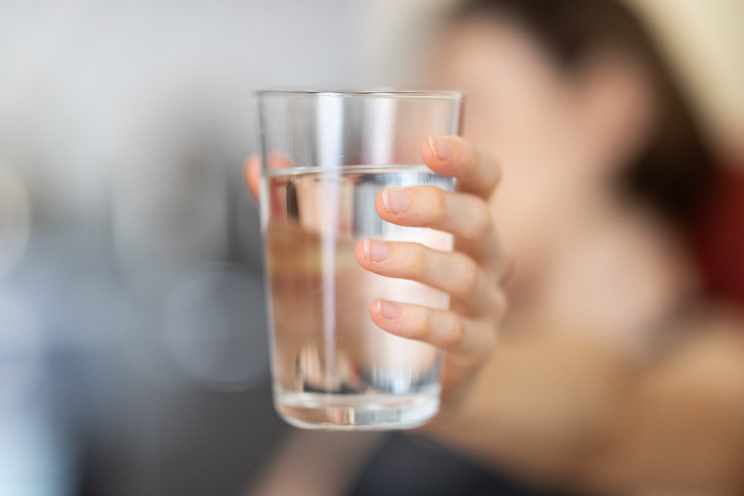 a hand in the foreground holding a glass of water. The person in the background is out of focus; How To Boost Your Daily Routine for a More Fulfilling Life