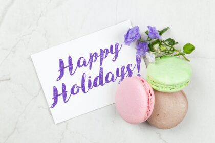 three macarons (green, pink, purple) are piled neatly on a table with a ourple flower. There is a piece of paper that says Happy Holidays! in a purple font; Festive Fables Imaginative Ways to Get Kids Excited for Every Holiday