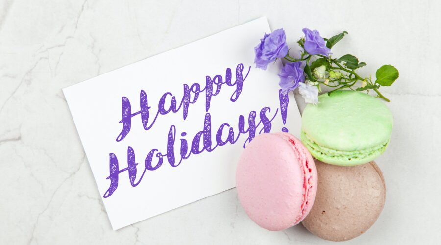 three macarons (green, pink, purple) are piled neatly on a table with a ourple flower. There is a piece of paper that says Happy Holidays! in a purple font; Festive Fables Imaginative Ways to Get Kids Excited for Every Holiday