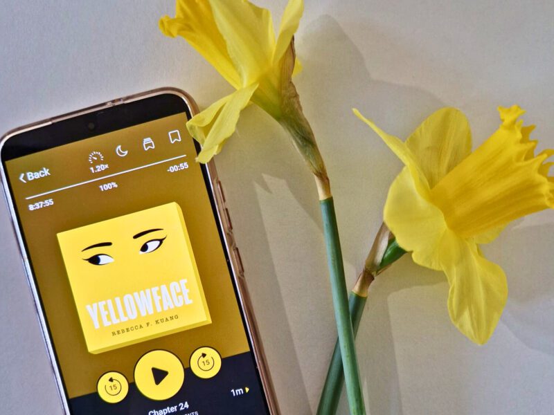 Yellowface by Rebecca F Kuang. The image shows a a cell phone laid flat on a white surface. On the phone is the audiobook. Nest to the phone are two single yellow daffodils laid across each other.