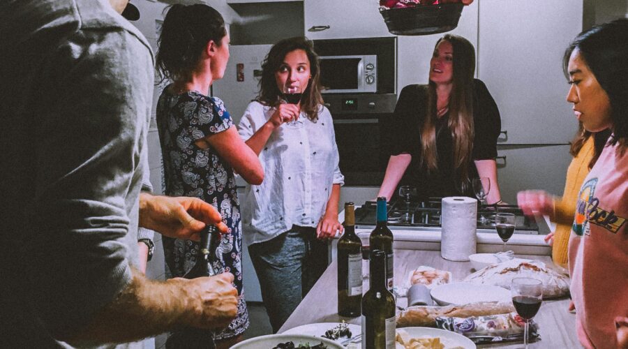 image shows a group of adults standing around a table laden with food and drinks; 6 Ways to Entertain Guests on a Budget