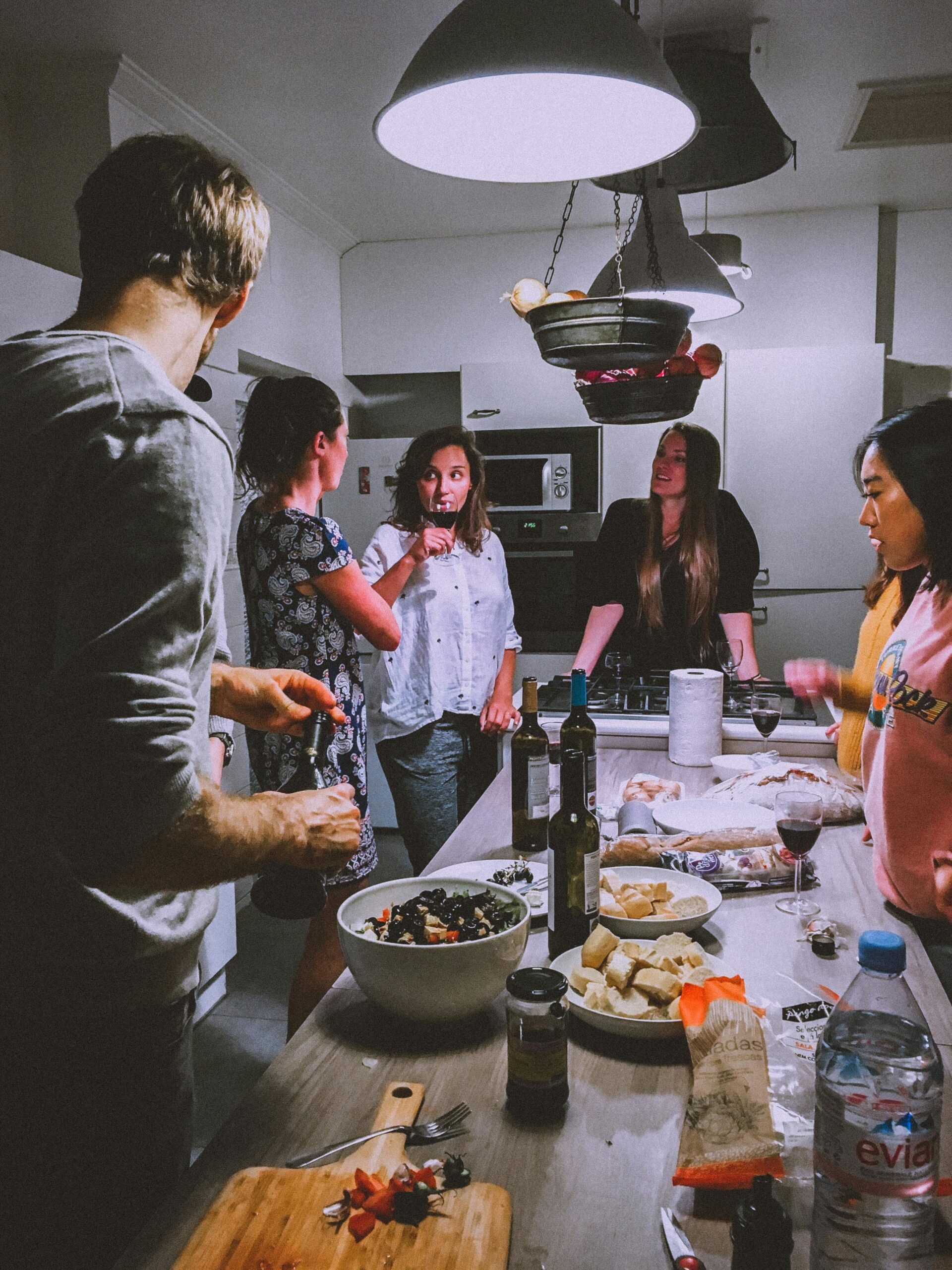 image shows a group of adults standing around a table laden with food and drinks; 6 Ways to Entertain Guests on a Budget