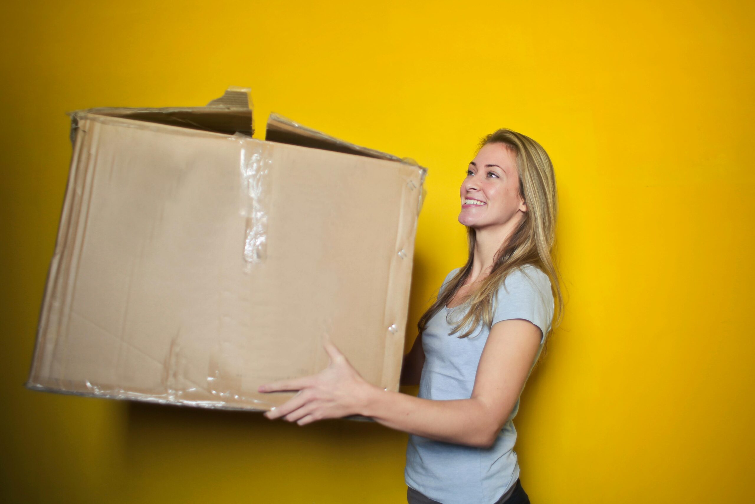 a woman in a grey shirt holding a brown cardboard box in front of a yellow wall; Classic Mistakes To Avoid When Moving To A New City