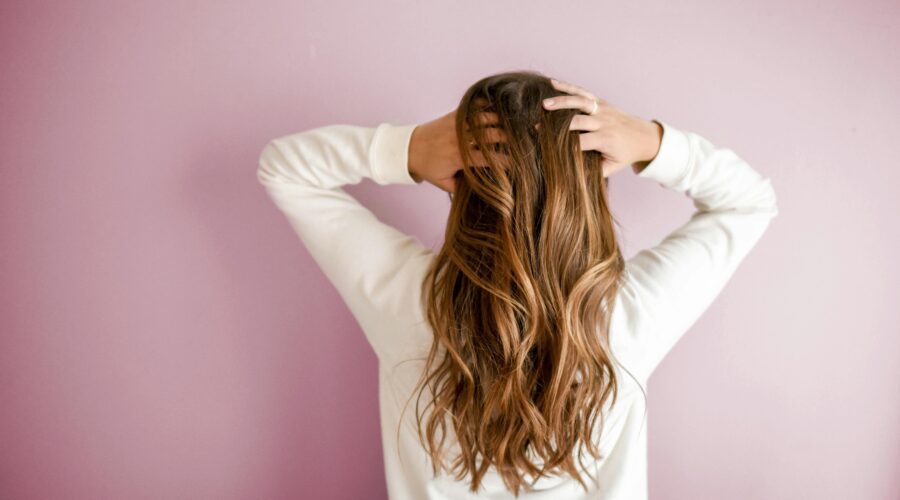 A woman wearing a white top with her back to the camera, holding her long light brown hair. she is standing against a light pink background