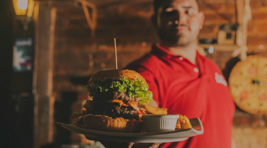 a man in a red shirt out of focus in the background, holding a burger in the foreground; What A New Restaurant Can Do To Make Their Diners Fall In Love