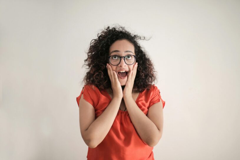 A woman with glasses, dark curly hair, and wearing an orange shirt holding her face and looking surprised