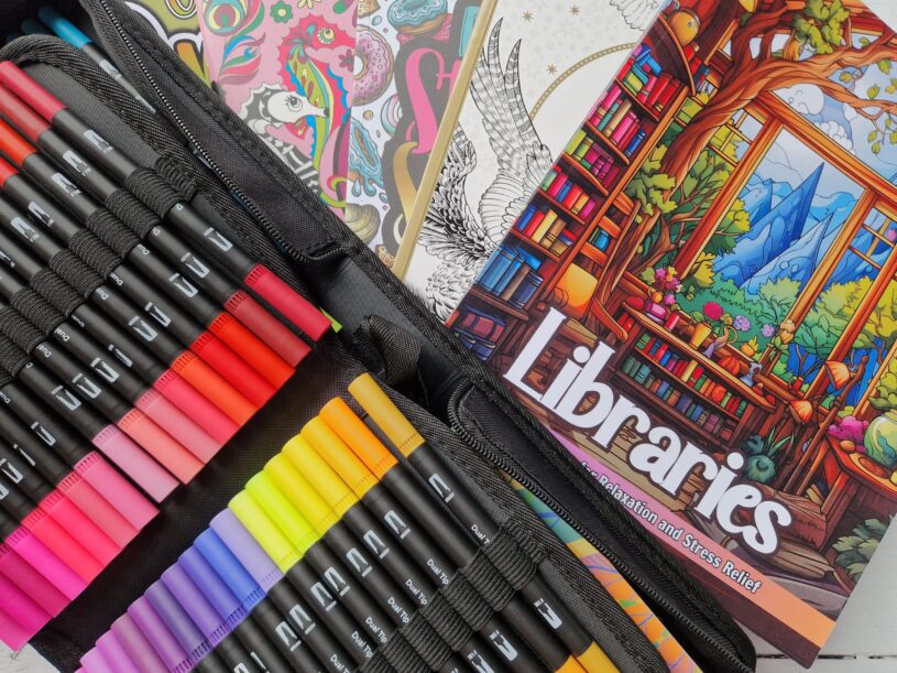 A view from above of a pile of adult colouring books and marker pens. The most prominent book is one that says "Libraries" on the front