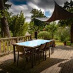 At the foreground of the image is a large brown deck area. On the deck is a long, narrow brown table with chairs around it. To the right of the image is a swing bench. The deck area has a brown canopy above it at the far end. In the background of the image are trees and bushes ; Creating the Perfect Outdoor Entertainment Space