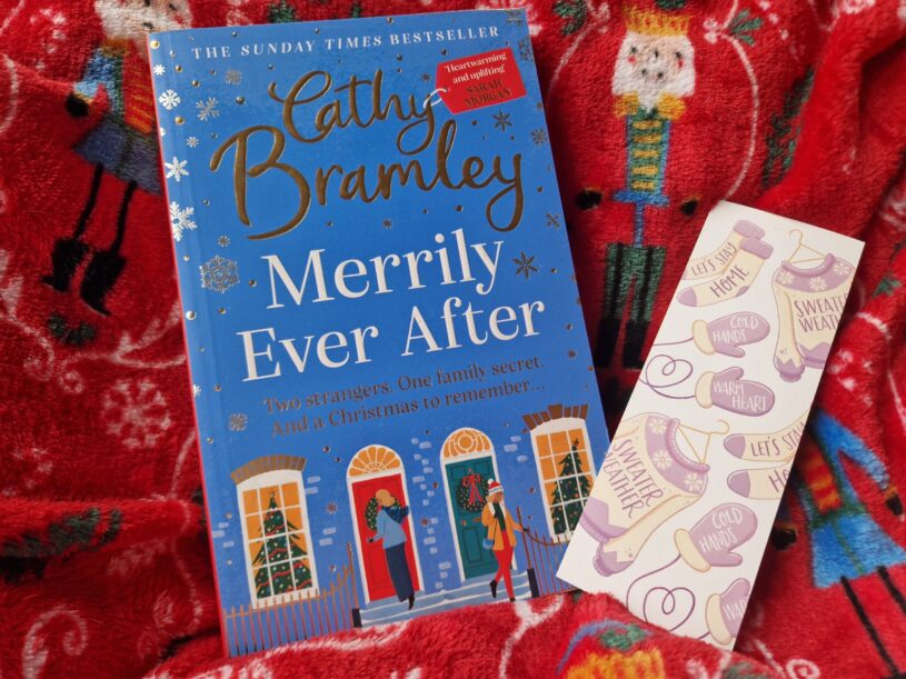 A paperback of Merrily Ever After by Cathy Bramley with a winter theme bookmark alongside it. The book is laid on a nutcracker print blanket