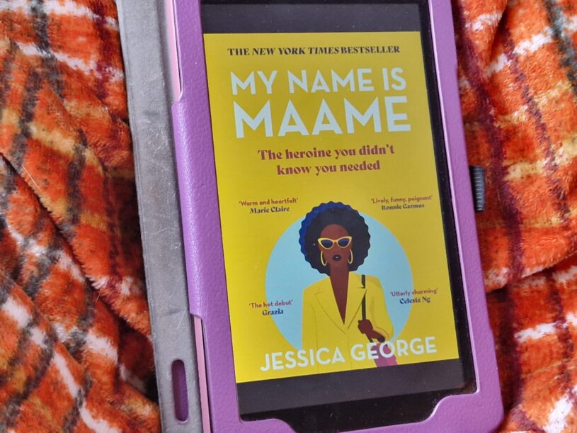 A Kindle fire device displaying the cover of My Name Is Maame by Jessica George. The device is laid on an orange checked blanket