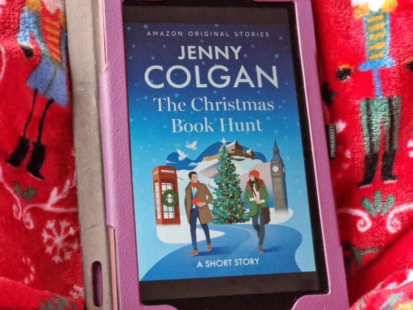 A Kindle fire device displaying the cover of The Christmas Book Hunt by Jenny Colgan. The device is laid on a nutcracker print blanket