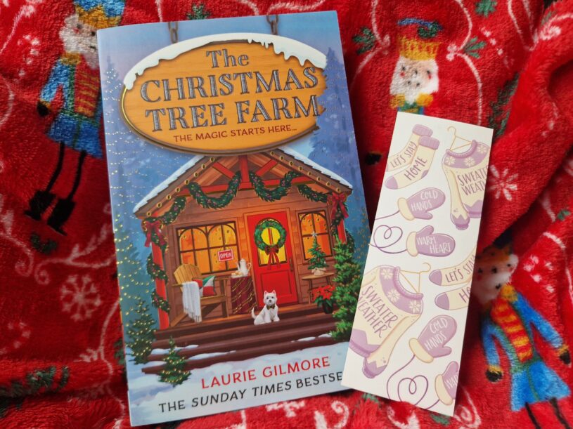 A paperback of The Christmas Tree Farm by Laurie Gilmore with a winter theme bookmark alongside it. The book is laid on a nutcracker print blanket