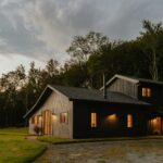 A large black house sitting on top of a green field. The house is surrounded by land and lots of trees behind it. There are clouds in the sky and the lights glow orange. Buying Your First Rural Property