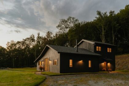 A large black house sitting on top of a green field. The house is surrounded by land and lots of trees behind it. There are clouds in the sky and the lights glow orange. Buying Your First Rural Property