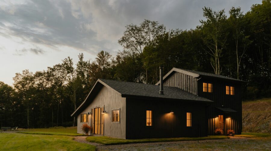 A large black house sitting on top of a green field. The house is surrounded by land and lots of trees behind it. There are clouds in the sky and the lights glow orange. Buying Your First Rural Property