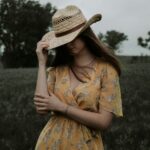 A woman with loose brunette hair stood in a field wearing a yellow floral dress. She is wearing a straw hat with her head facing down. One hand is holding the hat and the other is on the opposite elbow. Summer Wardrobe Tips for Country Living