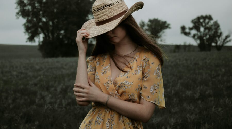 A woman with loose brunette hair stood in a field wearing a yellow floral dress. She is wearing a straw hat with her head facing down. One hand is holding the hat and the other is on the opposite elbow. Summer Wardrobe Tips for Country Living