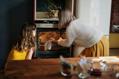 A woman in a white sweater and yellow skirt stood alongside a small girl in a yellow dress, in front of an oven putting a cake in it to bake; How to Choose the Right Oven for Your Kitchen