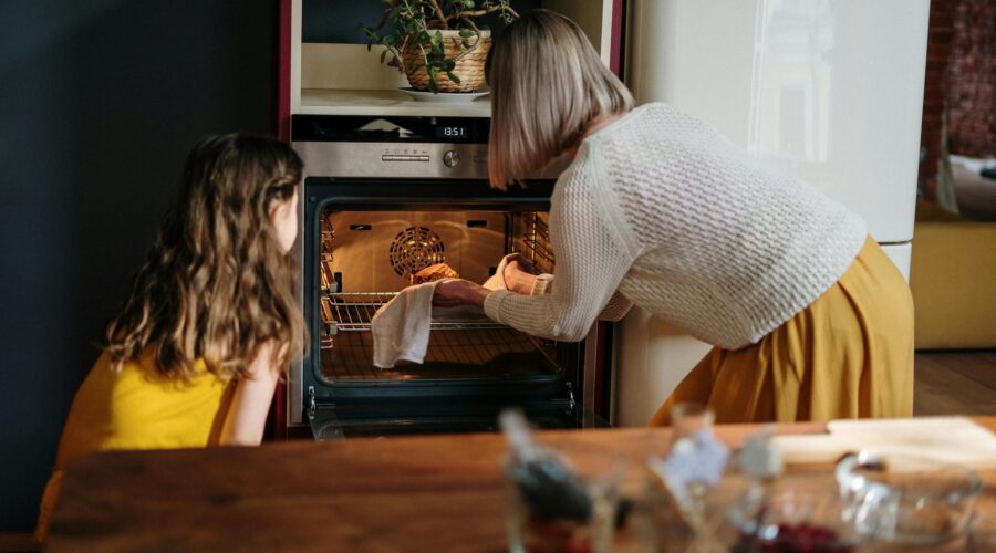 A woman in a white sweater and yellow skirt stood alongside a small girl in a yellow dress, in front of an oven putting a cake in it to bake; How to Choose the Right Oven for Your Kitchen